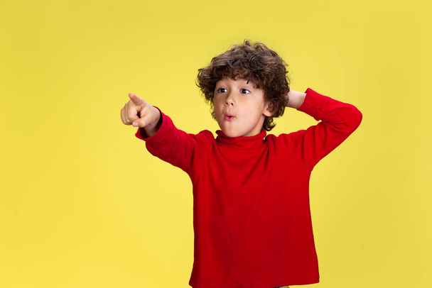 Pretty young curly boy in red wear on yellow studio background. Childhood, expression, fun. - Foto, Imagem