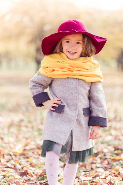 Belle petite fille dans un manteau gris, une écharpe jaune et un chapeau bordeaux posant pour la caméra dans un parc d'automne. Photo verticale - Photo, image