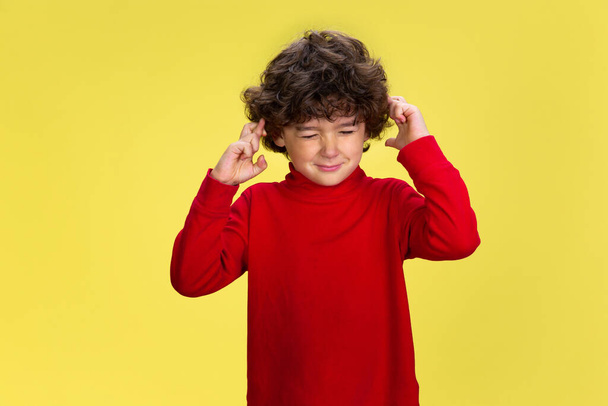 Pretty young curly boy in red wear on yellow studio background. Childhood, expression, fun. - Foto, afbeelding