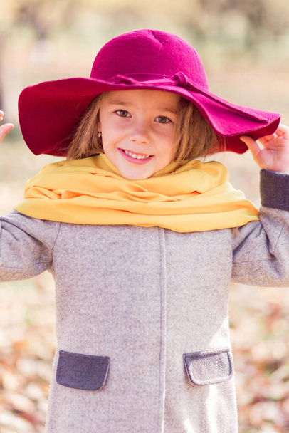 Beautiful little girl in a gray coat, a yellow scarf and a burgundy hat posing for the camera in an autumn park. Vertical photo - Photo, Image