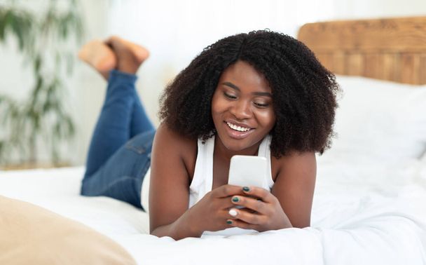 Felice ragazza nera sdraiata sul letto, utilizzando il telefono cellulare - Foto, immagini