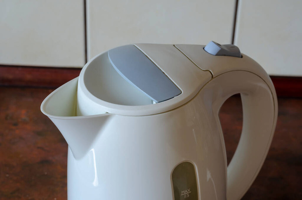 Weißer Wasserkocher auf dem Küchentisch. Eine Kunststoffvorrichtung zum Erhitzen von Wasser auf einem kastanienbraunen Tisch. Blick von oben schräg. Selektiver Fokus. - Foto, Bild