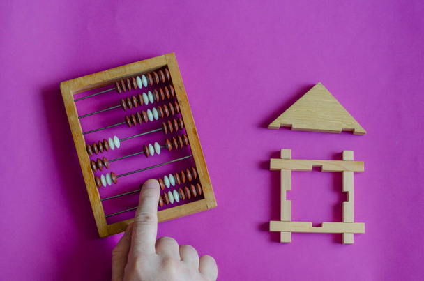 Mannelijke hand berekent een abacus naast speelgoed houten huis op lila achtergrond. Het begrip hypotheek of lening gedekt door onroerend goed. Uitzicht van bovenaf. - Foto, afbeelding
