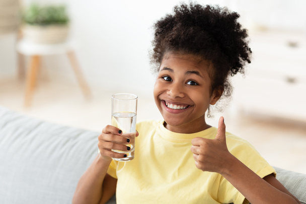 Afrikaans tiener meisje Holding glas van water Gesturing duimen-up Indoor - Foto, afbeelding