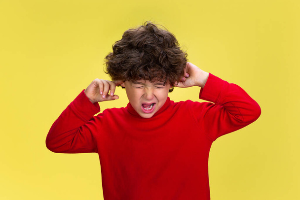 Pretty young curly boy in red wear on yellow studio background. Childhood, expression, fun. - Fotó, kép
