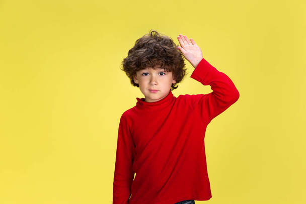 Pretty young curly boy in red wear on yellow studio background. Childhood, expression, fun. - Foto, Bild