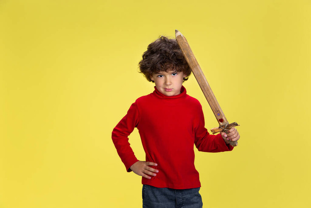 Mooie jonge krullende jongen in rood dragen op gele studio achtergrond. Jeugd, expressie, plezier. - Foto, afbeelding
