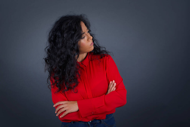 Image of upset pretty young brunette woman with curly hair wearing red shirt standing indoors with arms crossed. Looking with disappointed expression aside after listening to bad news.  - 写真・画像