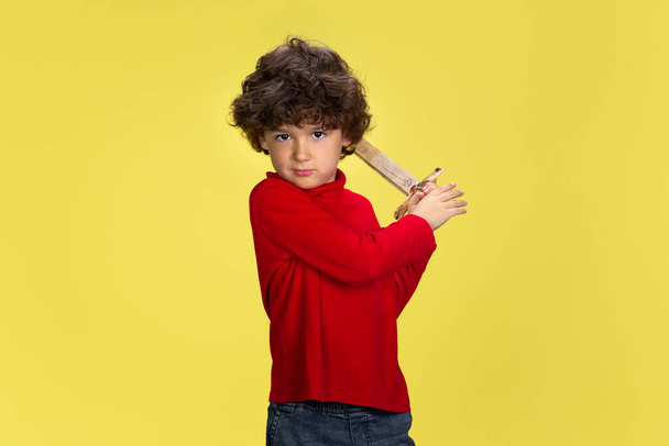 Ładny młody kręcony chłopiec w czerwonym stroju na żółtym tle studio. Dzieciństwo, ekspresja, zabawa. - Zdjęcie, obraz