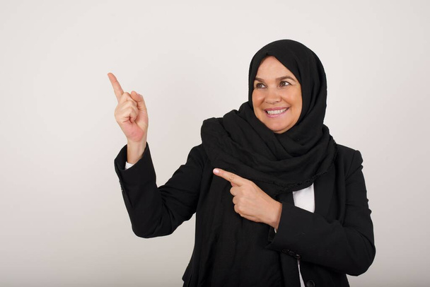 Красивая молодая мусульманка в черном хиджабе с позитивным выражением лица, одетая непринужденно, указывает носовым пальцем на пустое место для копирования рекламного текста или рекламы. Очаровательная женщина позирует одна - Фото, изображение
