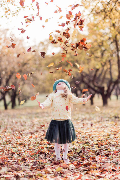 Красивая маленькая девочка в берете, желтом свитере и зеленой юбке из тюля в осеннем парке. Девушка бросает жёлтые листья. Вертикальное фото - Фото, изображение