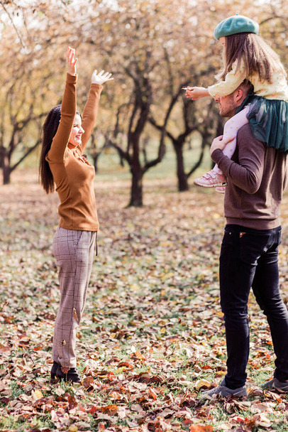 Mom, dad and their little daughter are walking in the autumn park. The girl sits on dad's shoulders. Mom throws autumn leaves up. Vertical photo - Foto, Imagem