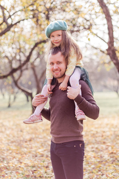 Papa en dochter lopen in het herfstpark. De dochter zit op de schouders van papa. Pa en dochter lachen en kijken naar de camera. Verticale foto - Foto, afbeelding
