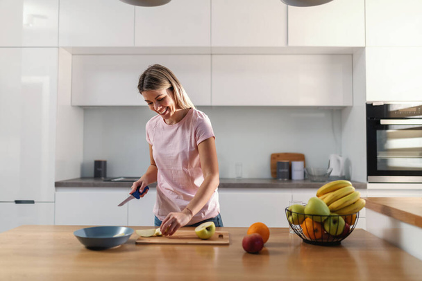 Güzel, gülümseyen sarışın kadın mutfakta duruyor ve meyve doğruyor. Corona virüsü sırasında vitaminler önemlidir.. - Fotoğraf, Görsel