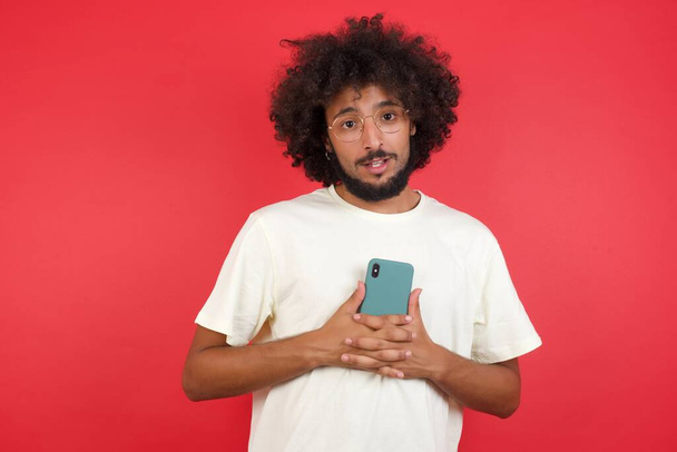 Afro saçlı genç adam kırmızı duvara akıllı telefon tutuyor.    - Fotoğraf, Görsel