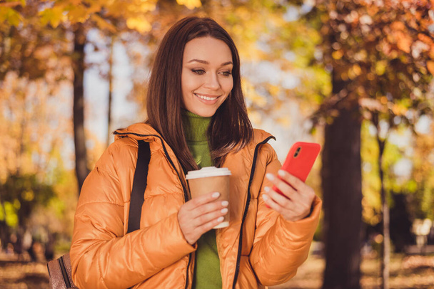 Photo of cute lovely lady smiling hand hold telephone look screen cup drink coffee post share recent photo autumn park wear green turtleneck sweater orange coat trousers outdoors - Foto, Imagen