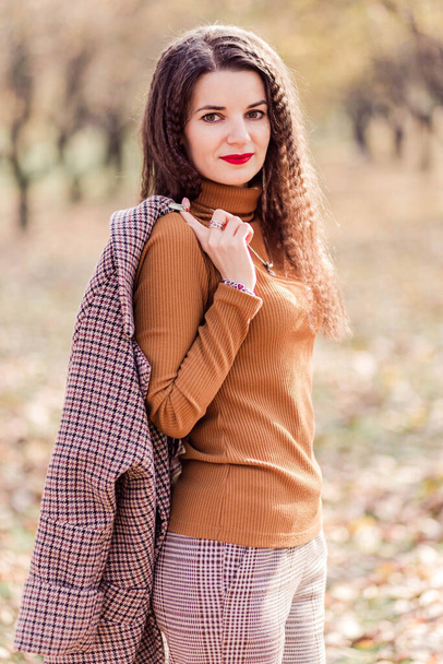 Beautiful brunette woman in trousers , turtleneck and coat over her shoulder posing in autumn park. Woman looks at the camera . Vertical photo - Photo, Image