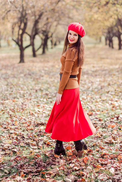 秋の公園でポーズをとっている赤いベレー帽、茶色のタートルネックと赤いスカートの美しいブルネットの女性。垂直写真 - 写真・画像