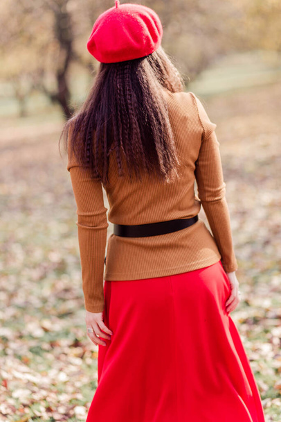 秋の公園でポーズをとっている赤いベレー帽、茶色のタートルネックと赤いスカートの美しいブルネットの女性。女の子はカメラに背を向けた縦の写真 - 写真・画像