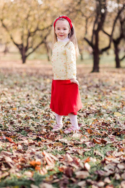 Malá holčička v dlouhé červené sukni, teplé žluté bundě a červeném obvazu na hlavě bzučí sama v podzimním parku. Dívka se otočí, usměje se a podívá se do kamery. Svislá fotografie - Fotografie, Obrázek