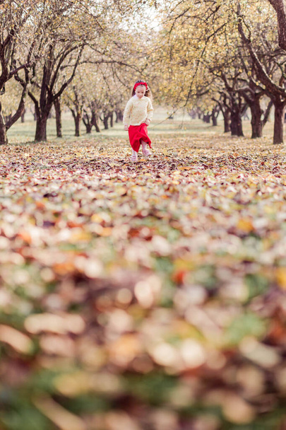 Een klein meisje in een lange rode rok, een warm geel jasje en een rood verband om haar hoofd zoemt alleen in het herfstpark. Het meisje heeft plezier en rent door het park. Verticale foto - Foto, afbeelding