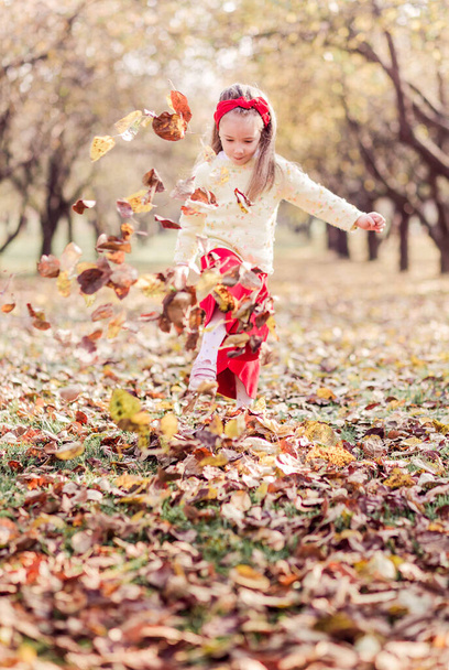 Een klein meisje in een lange rode rok, een warm geel jasje en een rood verband om haar hoofd zoemt alleen in het herfstpark. Het meisje speelt en gooit de bladeren met haar voeten. Verticale foto - Foto, afbeelding