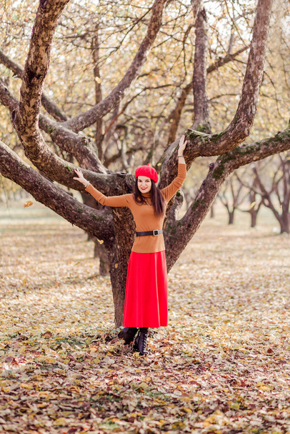 Güz parkında poz veren kırmızı bereli, kahverengi boğazlı ve kırmızı etekli güzel esmer kadın. Uzun bir ağacın yanında poz veren kız. Dikey fotoğraf - Fotoğraf, Görsel