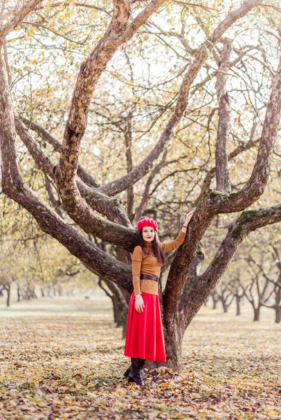 Gyönyörű barna nő piros svájcisapkában, barna garbóval és piros szoknyával pózol egy őszi parkban. Egy lány pózol egy magas fa mellett. Függőleges fénykép - Fotó, kép