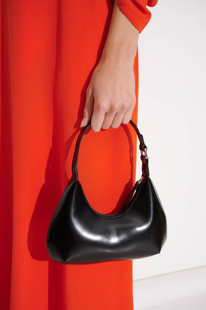 女の子の手の中に美しい黒い革のハンドバッグ。高級デザイナーのバッグに対して赤ドレス。ファッショナブルな現代的なアクセサリー。垂直写真. - 写真・画像