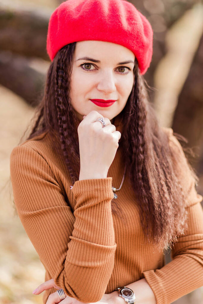 Krásná brunetka v červeném baretu, hnědý rolák pózující v podzimním parku. Dívka se dívá do kamery. Svislá fotografie - Fotografie, Obrázek