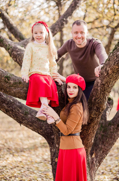 Mamá, papá e hija pasan tiempo juntos en el parque. Papá y su hija subieron a un árbol, Maa está de pie cerca. Familia mirando a la cámara. Foto vertical - Foto, imagen