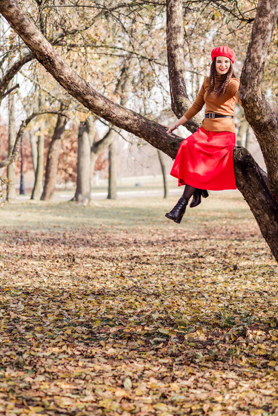 Όμορφη μελαχρινή γυναίκα με κόκκινο μπερέ, καφέ ζιβάγκο και κόκκινη φούστα κάθεται ψηλά σε ένα κλαδί δέντρου σε ένα φθινόπωρο πάρκο. Κάθετη φωτογραφία - Φωτογραφία, εικόνα