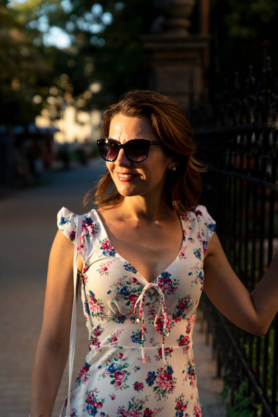 Beautiful woman in flower dress wearing sunglasses at sunset. Beauty and Fashion luxury style. Golden hour photo - Foto, Imagem