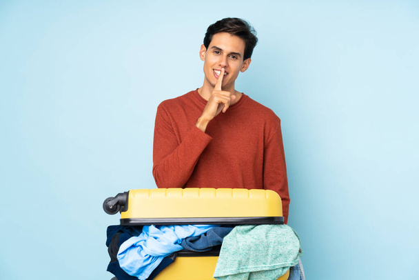 Мужчина с чемоданом, полным одежды на изолированном синем фоне делает жест молчания - Фото, изображение