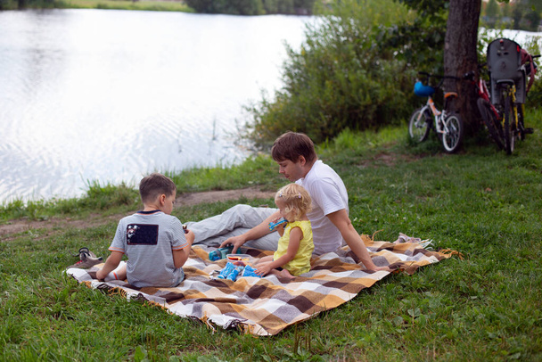 Πατέρας και παιδιά κάνουν πικ-νικ κοντά στη λίμνη. Η οικογένεια κάθεται στο καρό και τρώει. Ποδήλατα στέκονται κοντά - Φωτογραφία, εικόνα