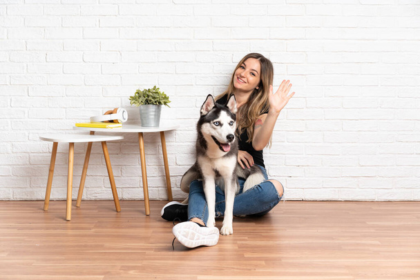 Νεαρή όμορφη γυναίκα με το γεροδεμένο σκυλί της να κάθεται στο πάτωμα σε εσωτερικούς χώρους χαιρετώντας με το χέρι με χαρούμενη έκφραση - Φωτογραφία, εικόνα