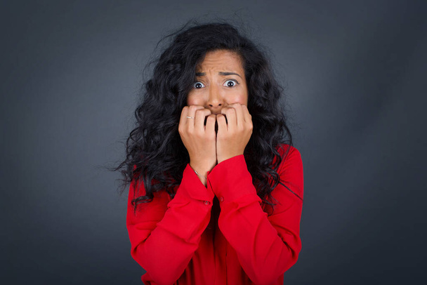 beautiful young brunette woman with curly hair wearing red shirt covering her mouth with her hands and standing outdoors. Scared from something or someone and biting nails. - Photo, Image
