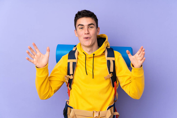 Teenager caucasian mountaineer man with a big backpack isolated on purple background with surprise facial expression - Foto, afbeelding