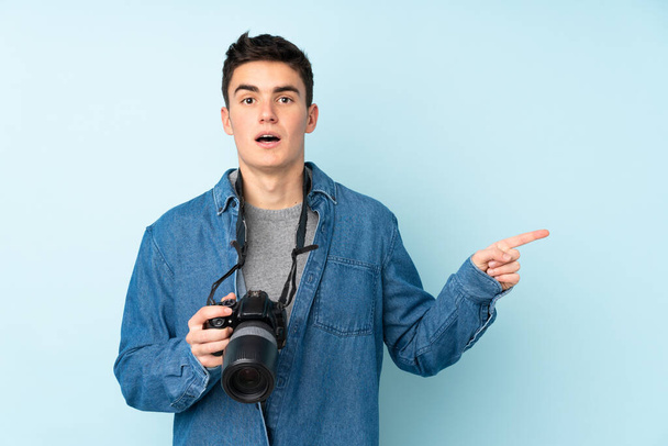 Teini valokuvaaja mies eristetty sinisellä taustalla yllättynyt ja osoittaa sormella sivulle - Valokuva, kuva