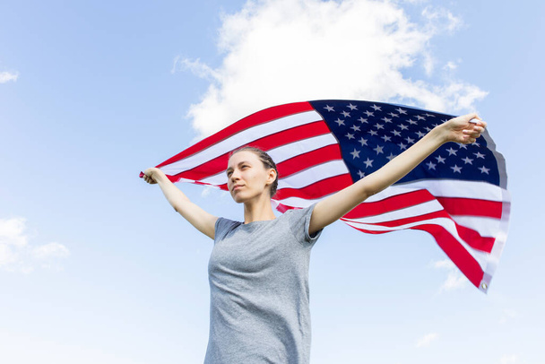 Mujer joven con una cara seria de expresión sosteniendo una bandera estadounidense en protesta. Demostración. Bandera de EE.UU. contra el cielo. - Foto, imagen