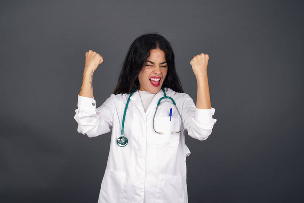 Valkoihoinen brunette lääkäri nainen yllään lääketieteellinen univormu ja stetoskooppi, iloitsee menestyksestä ja voitosta puristamalla nyrkkejään ilolla saavuttaakseen tavoitteensa ja tavoitteensa.. - Valokuva, kuva
