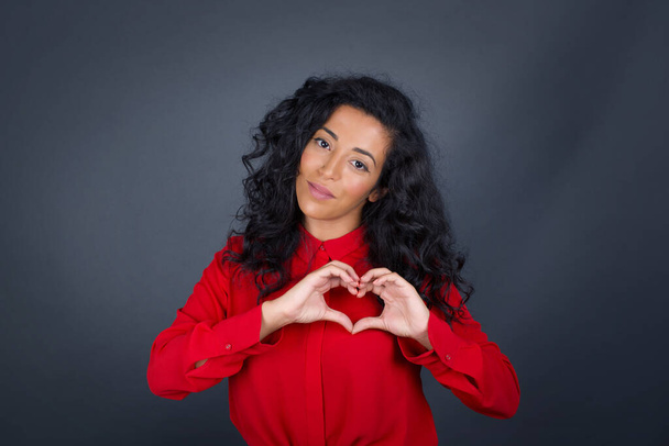Beautiful brunette woman with curly hair wearing red shirt over gray background smiling in love and showing heart symbol and shape with hands. Romantic concept. - Foto, imagen