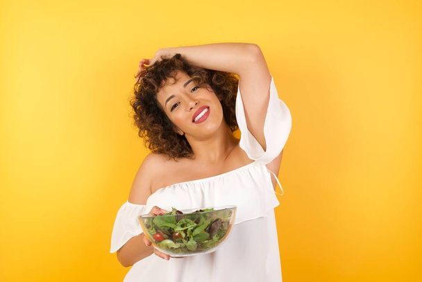 Portrait of beautiful brunette female holding a salad dressed casual looks with excitement at camera, keeps hands raised over head notices something unexpected, isolated over yellow wall. Lovely woman reacts on sudden news. - Фото, изображение