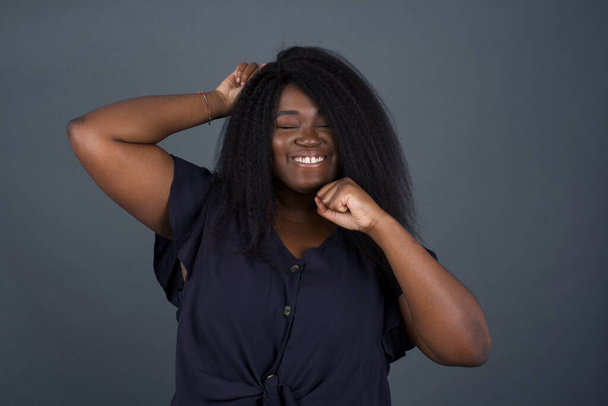 Attractive young African American  woman celebrating a victory punching the air with her fists and a beaming toothy smile over a gray studio background with copy space - Foto, Imagen