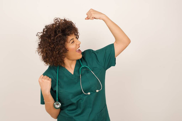 belle médecin arabe femme portant un uniforme médical célébrant une victoire frapper l'air avec ses poings et un sourire rayonnant de dents sur un fond studio bleu avec espace de copie. - Photo, image