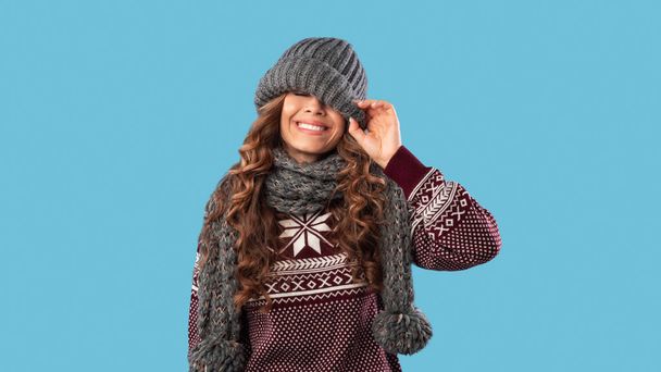 Zimní móda. Mladá dívka stahuje teplou čepici a usmívá se přes modré pozadí - Fotografie, Obrázek