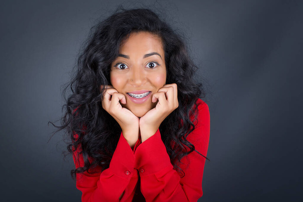 Dreamy young brunette woman with curly hair wearing red shirt keeps hands pressed together under chin, looks with happy expression, has toothy smile, isolated over gray background - 写真・画像