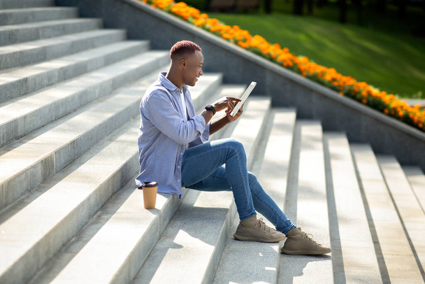 Πλευρική άποψη του χαρούμενου μαύρου άντρα χρησιμοποιώντας το tablet υπολογιστή σε πέτρινες σκάλες στο πάρκο της πόλης - Φωτογραφία, εικόνα