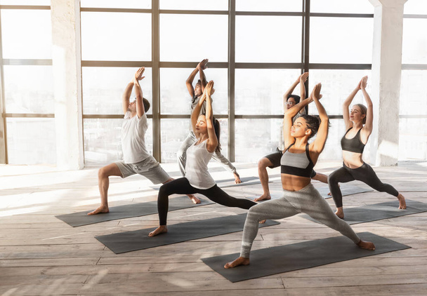 Group Of Young Yoga Enthusiasts Training Together In Studio, Practicing Warrior Pose - Photo, image