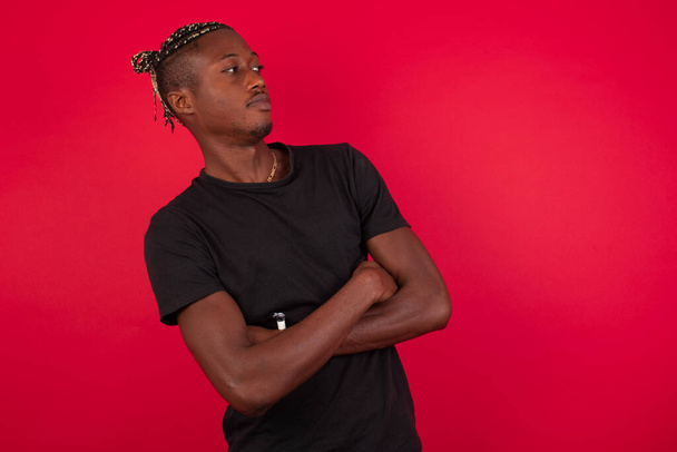 Image of upset young African American man wearing black t-shirt over red background standing indoors with arms crossed. Looking with disappointed expression aside after listening to bad news. Confident young man. - Φωτογραφία, εικόνα